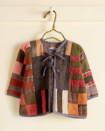 The Oak Chest Lined Baby Jacket - OS, a multicolored patchwork quilt jacket crafted from an antique wool quilt with hand-mending details, hangs on a green wire hanger against a beige wall.