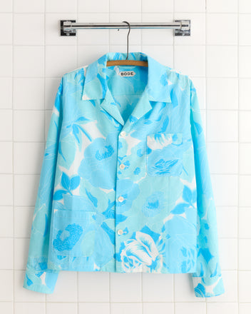 The Ocean Florals Shirt - L/XL is a light blue button-up with a bright blue floral pattern, crafted from deadstock fabric, shown on a hanger against a white tiled wall.
