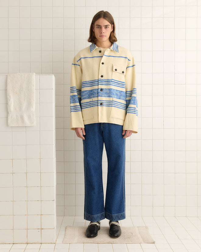 A person wearing the Oceanside Stripe Blanket - L/XL stands on a tiled floor next to a towel, showcasing their unique style with its cream and blue-striped design paired with wide-leg jeans.