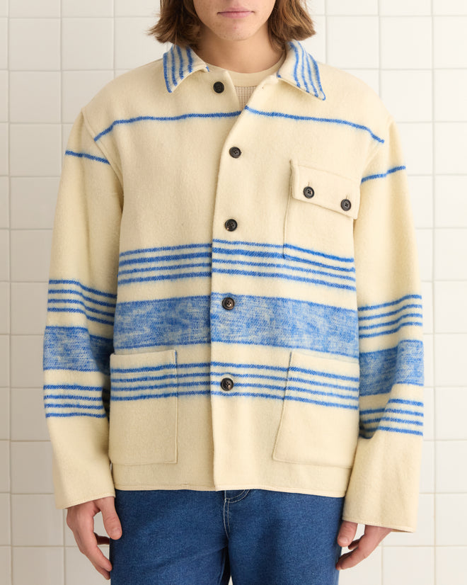 A person wears an Oceanside Stripe Blanket in L/XL, featuring a unique cream base with blue stripes, paired with jeans against a tiled backdrop.