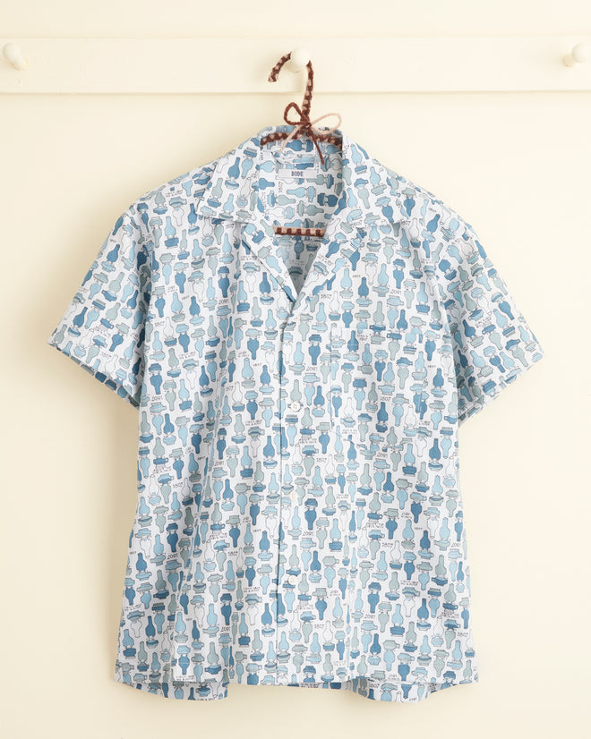 A unique button-up shirt called the Oil Lamp Shirt - XS/S, featuring a distinct blue and white pattern with silhouettes of various bottle shapes, crafted from special textile, hanging on a white wall hook.