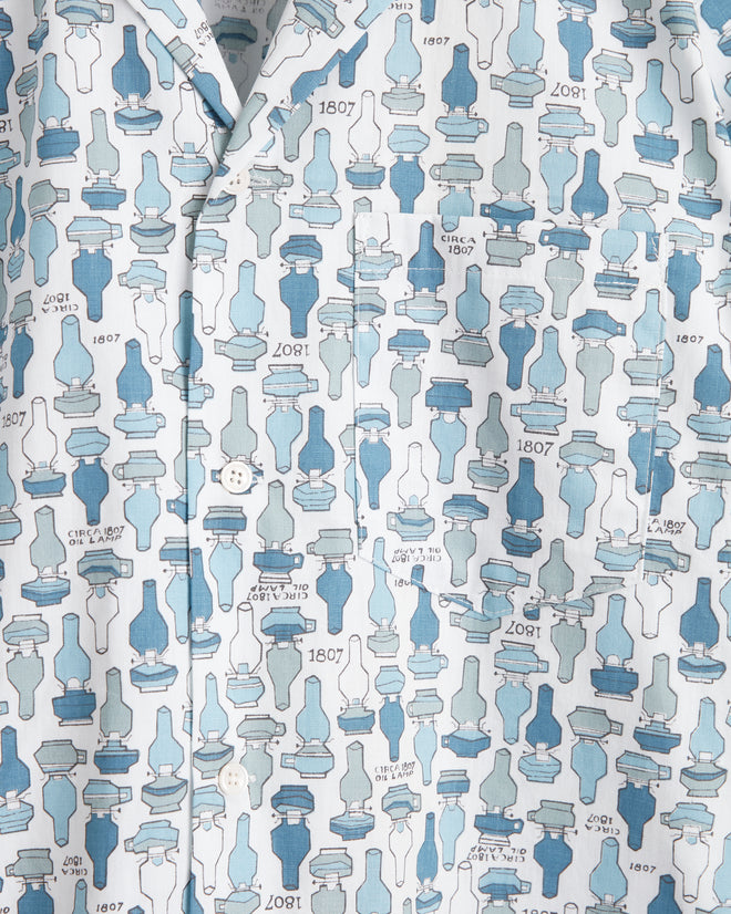 A unique button-up shirt called the Oil Lamp Shirt - XS/S, featuring a distinct blue and white pattern with silhouettes of various bottle shapes, crafted from special textile, hanging on a white wall hook.