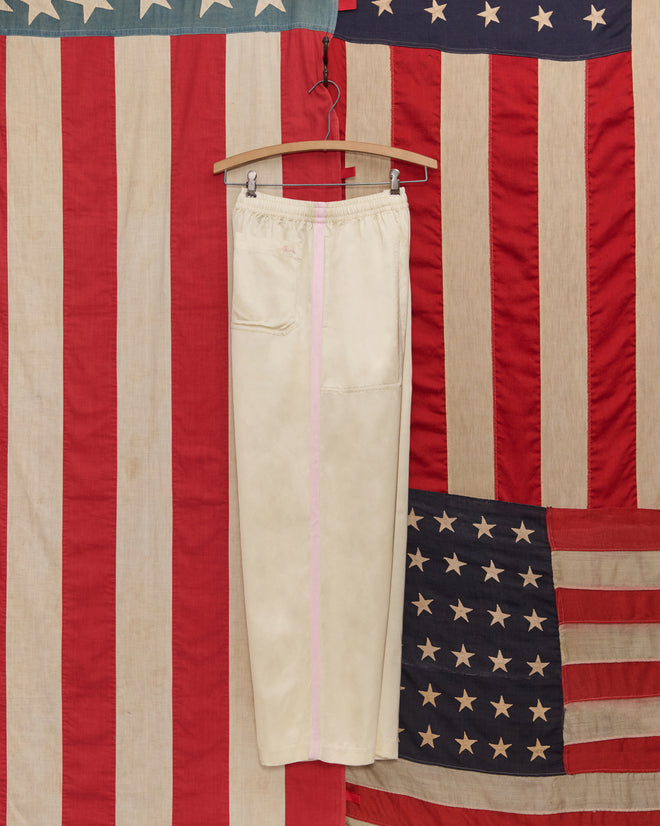 The Satin Track Pants in White/Pink, featuring a pink side stripe, hang on a wooden hanger against an American flag backdrop.