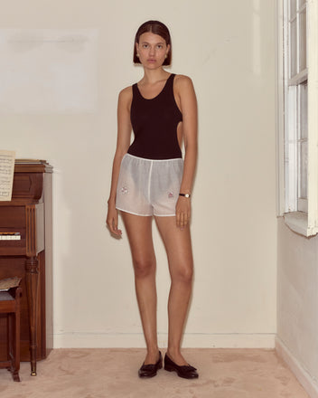 A woman in a black tank top and On Air Tap Shorts - White stands by a piano in a sunlit, minimally furnished room.