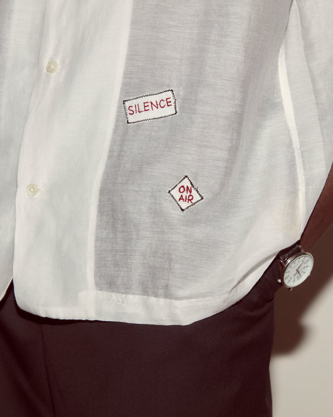 The On Air Short Sleeve is a white cotton-silk shirt with embroidered "SILENCE" and "ON AIR" patches, inspired by recording studio shirts. Pair it with dark pants and a watch for a modern look.