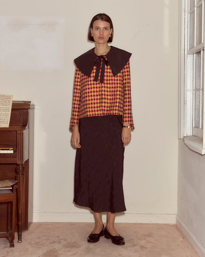 Woman in the Orana Collar Jacket, styled with a black skirt and flats in 1960s French fashion, stands indoors by a piano.
