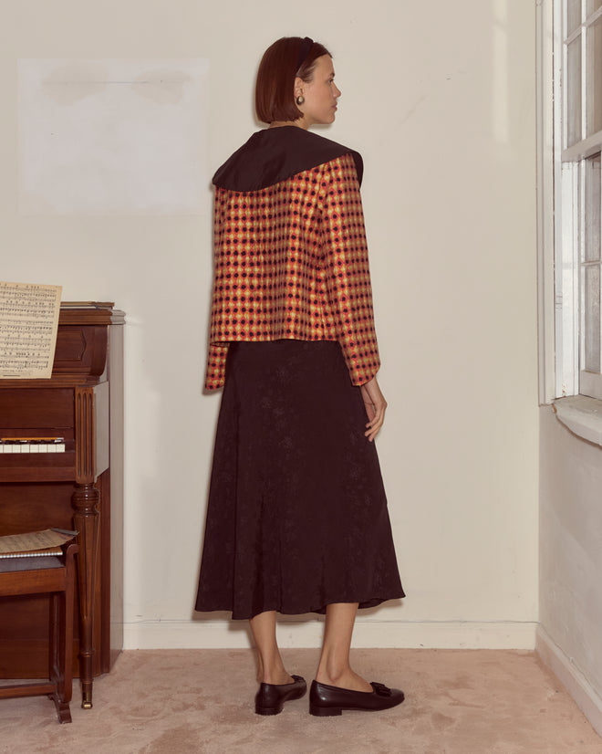 Woman in an Orana Collar Jacket and black skirt stands indoors by a piano and window, facing sideways.