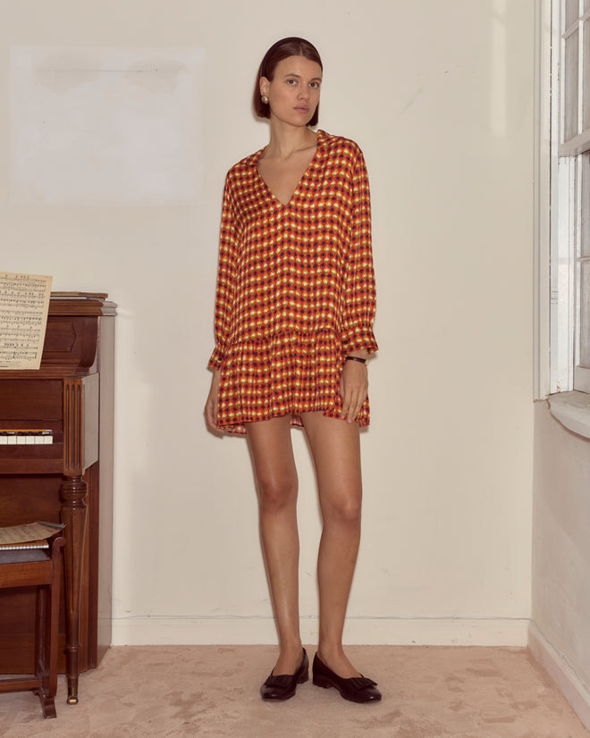 A woman wearing the Orana Dress, featuring a 1960s-inspired French checkered pattern, stands indoors beside a piano and near a sunlit window.