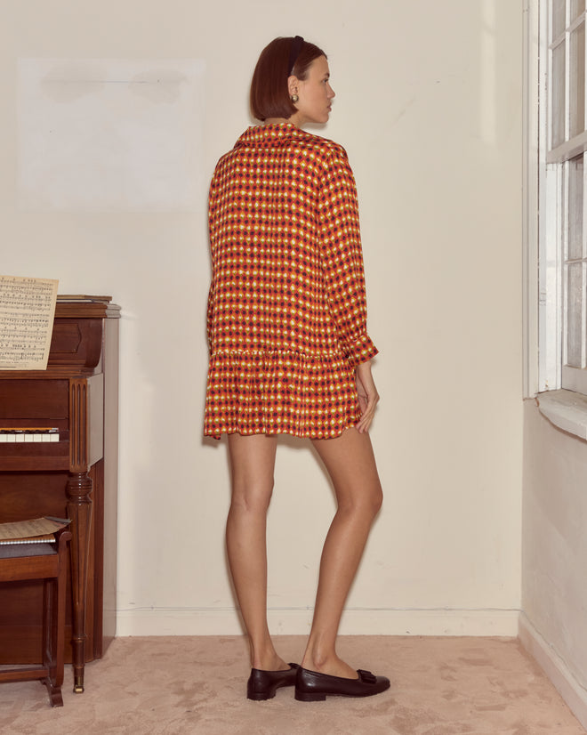A woman stands by a piano and window, facing sideways, wearing the Orana Dress—a scarlet silk drop-waist design inspired by 1960s French patterns.