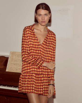 A woman in the Orana Dress, a scarlet silk drop-waist dress inspired by 1960s French patterns, stands before a piano with sheet music.