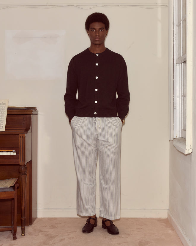 A man stands indoors next to a piano wearing the Orchard Cardigan - Black, striped pants, and dark shoes.