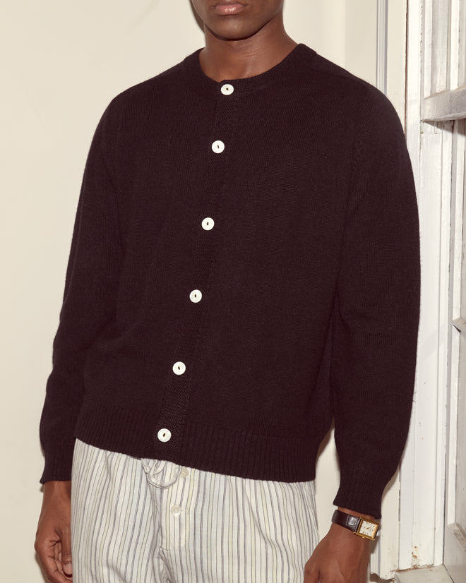 Man wearing the Orchard Cardigan - Black with white buttons and striped pants, standing near a window.