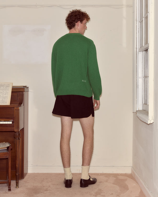A person in an Orchard Cardigan - Kelly Green and black shorts stands facing a cream-colored wall near a piano and window.