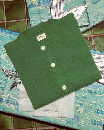 The Orchard Cardigan in Kelly Green, made from merino wool, is folded neatly atop blue patterned wrapping paper decorated with silver leaves.