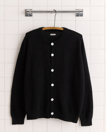 The Orchard Cardigan - Black, a merino wool cardigan with white buttons, is displayed on a wooden hanger against a white tiled wall.