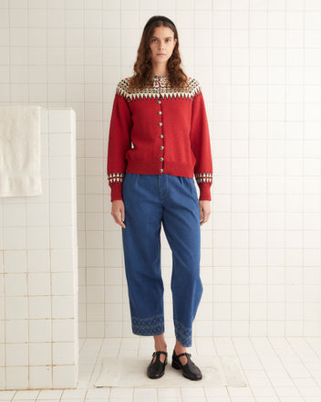 A person stands against a tiled background wearing the Oslo Cardigan in a red pattern made from a Norwegian wool blend, blue jeans with patterned cuffs, and black shoes.