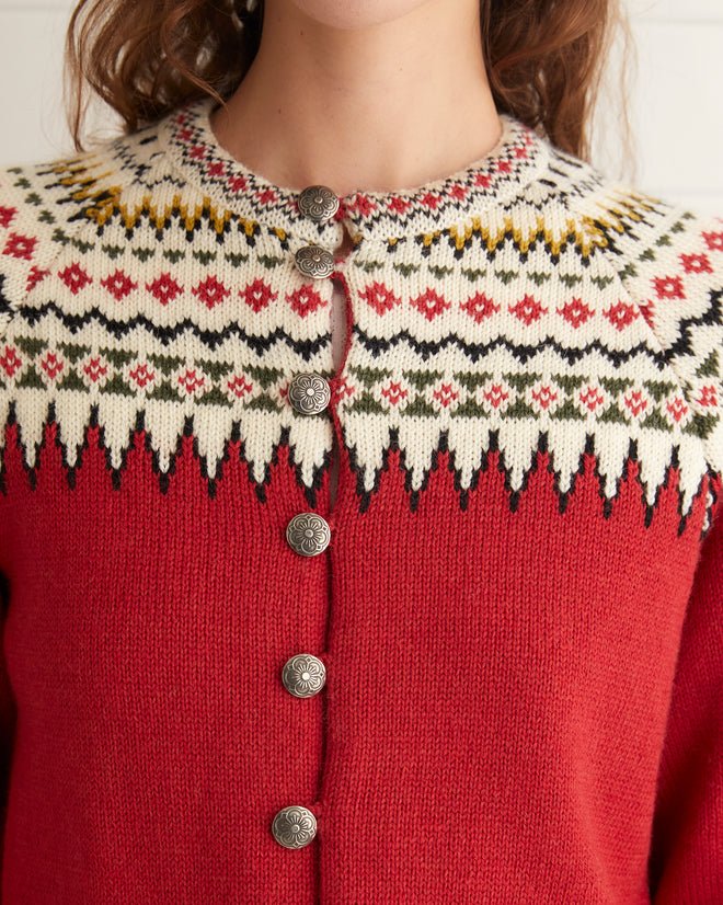 A person stands against a tiled background wearing the Oslo Cardigan in a red pattern made from a Norwegian wool blend, blue jeans with patterned cuffs, and black shoes.