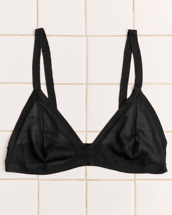 The Knit Bra - Black, featuring a vintage-inspired design with thin straps and mesh detailing, is displayed laid flat on a white tiled surface.