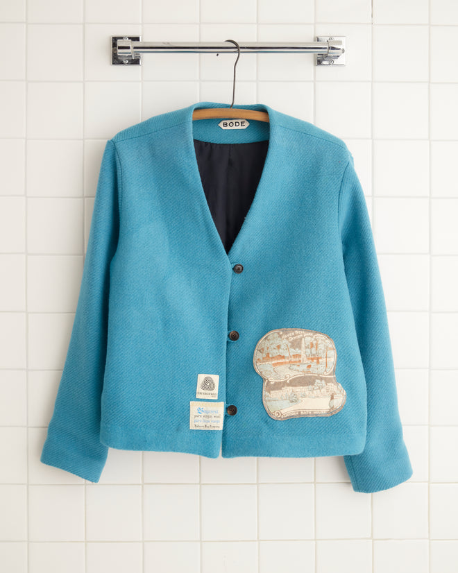 The Pacific Wool Jacket - 2, a midcentury-style garment with a boxy fit, hangs on a tile wall. This wool jacket is enhanced with brown buttons and features a vintage patch and label on the front, adding classic charm to its design.