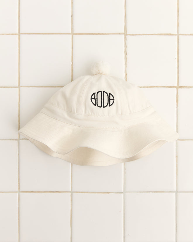 The Paneled Grandfather Hat - White, crafted from midweight canvas and featuring "BODE" embroidery, hangs on a tiled wall—ideal for your next college reunion.