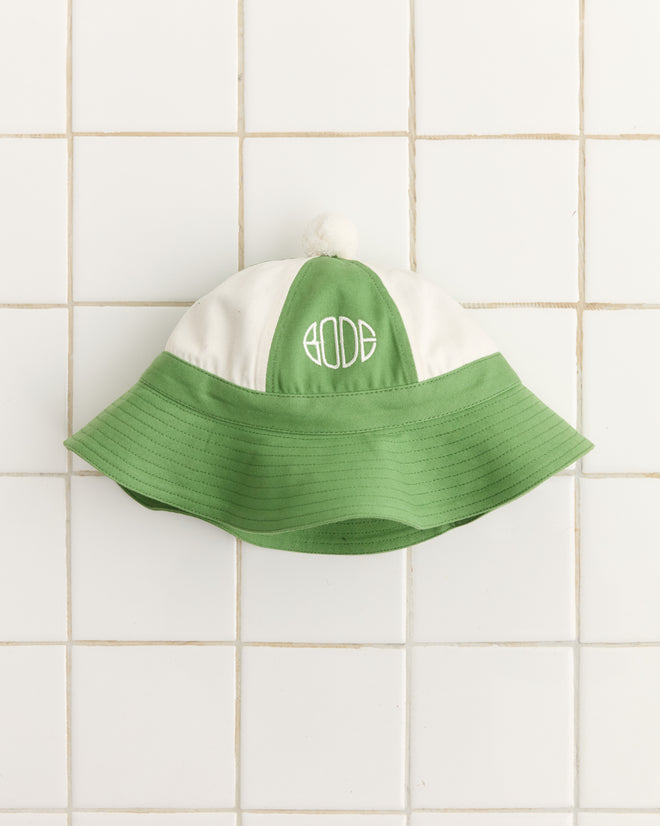 The Paneled Grandfather Hat in green/white, crafted from 100% cotton with Bode embroidery, is showcased on a tiled background. Ideal for that college reunion hat vibe.