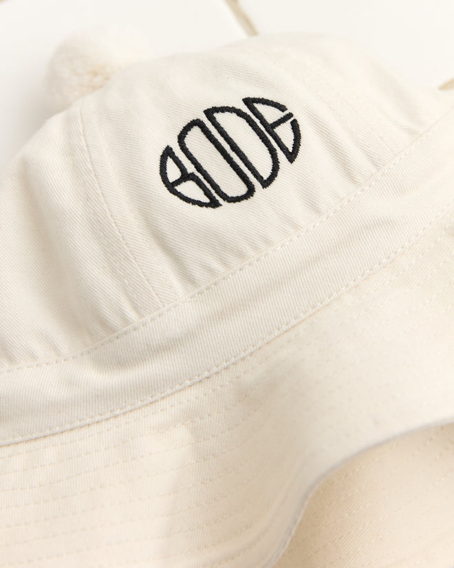 The Paneled Grandfather Hat in white features a pom-pom, Bode embroidery, and a black circular front logo, meticulously crafted from 100% cotton.