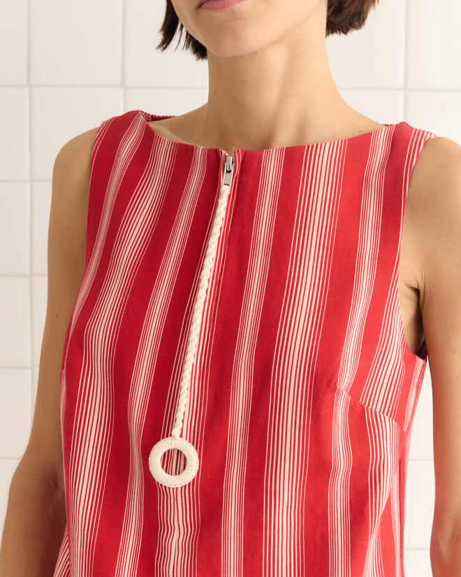 A person in a Parasol Stripe Dress with a large zipper pull, featuring bold red and white awning stripes, stands against a white tiled wall.