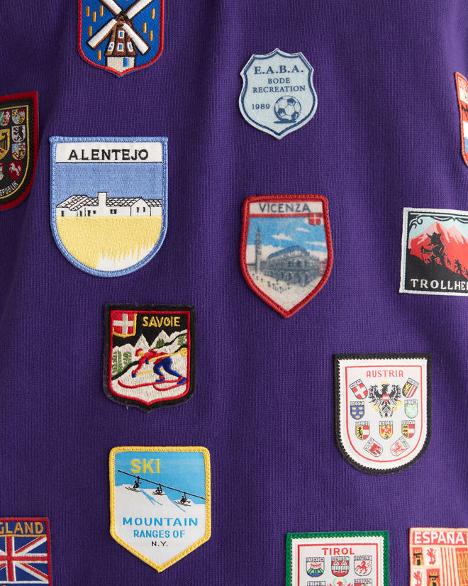 A man is standing in a tiled room clad in the Passport Patch Rugby Shirt in purple, featuring vintage patches, and black pants.