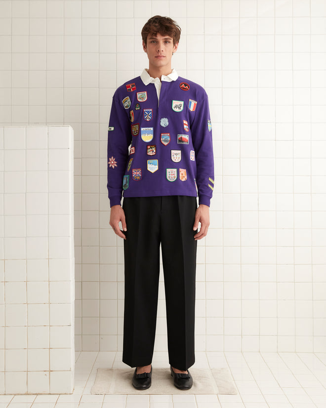 A man is standing in a tiled room clad in the Passport Patch Rugby Shirt in purple, featuring vintage patches, and black pants.