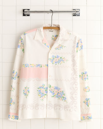 The Pastel Garland Shirt - S/M is a long-sleeve, button-up top with vintage tablecloth fabric featuring pastel floral and paisley motifs, displayed on a hanger against white tiles for a unique look.