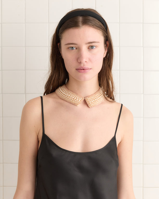 A woman in a black dress with the Pearl Collar stands against a tiled wall, wearing a black headband. Her look is completed with subtle silk accessories, evoking a 1960s reproduction style.
