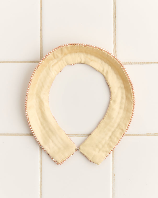 The Pearl Collar is a detachable beige accessory featuring pink and pearl beading, reminiscent of 1960s style, set against a white tiled background.