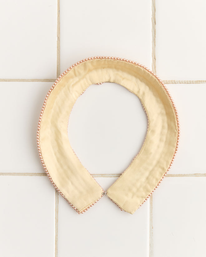 The Pearl Collar is a beige pearl-beaded accessory with an elegant pearl trim, beautifully displayed against a white tile background.