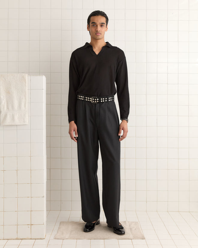 In a tiled room, a person in black sports the Clover Charm Belt - Black with vintage appeal. Their gleaming black shoes with heavyweight suede accents complement the edgy touch of their wet hair.