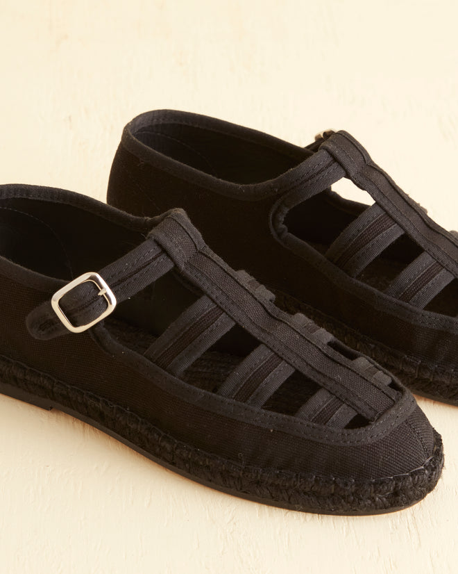 The Peggy Espadrilles - Black, featuring a pair of black, closed-toe espadrilles with a buckle strap, multiple cutouts, and a braided jute sole, placed on a light surface: the perfect summer shoes.