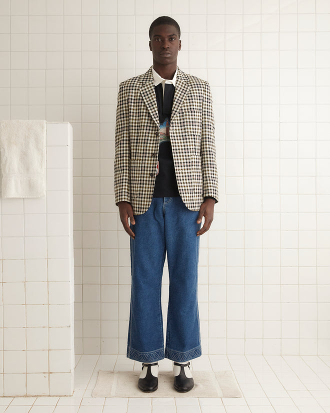 A person wearing a Pelican Plaid Suit Jacket - 42, paired with a graphic shirt and jeans, stands against a wooden wall.