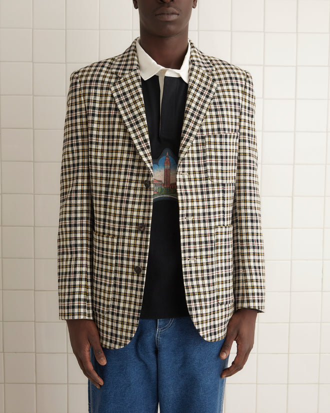 A person wearing a Pelican Plaid Suit Jacket - 42, paired with a graphic shirt and jeans, stands against a wooden wall.