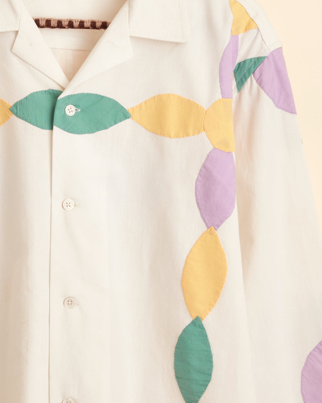 The Petal Appliqué Shirt - M/L, showcasing unique green, yellow, and purple leaf-like patterns reminiscent of a 1940s-1950s quilt top, hangs on a braided hanger against a beige background.