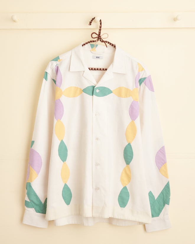 The Petal Appliqué Shirt - M/L, showcasing unique green, yellow, and purple leaf-like patterns reminiscent of a 1940s-1950s quilt top, hangs on a braided hanger against a beige background.