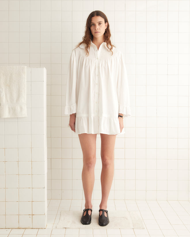 A person stands in a white tiled room wearing the Petaluma Dress in white, paired with black shoes.