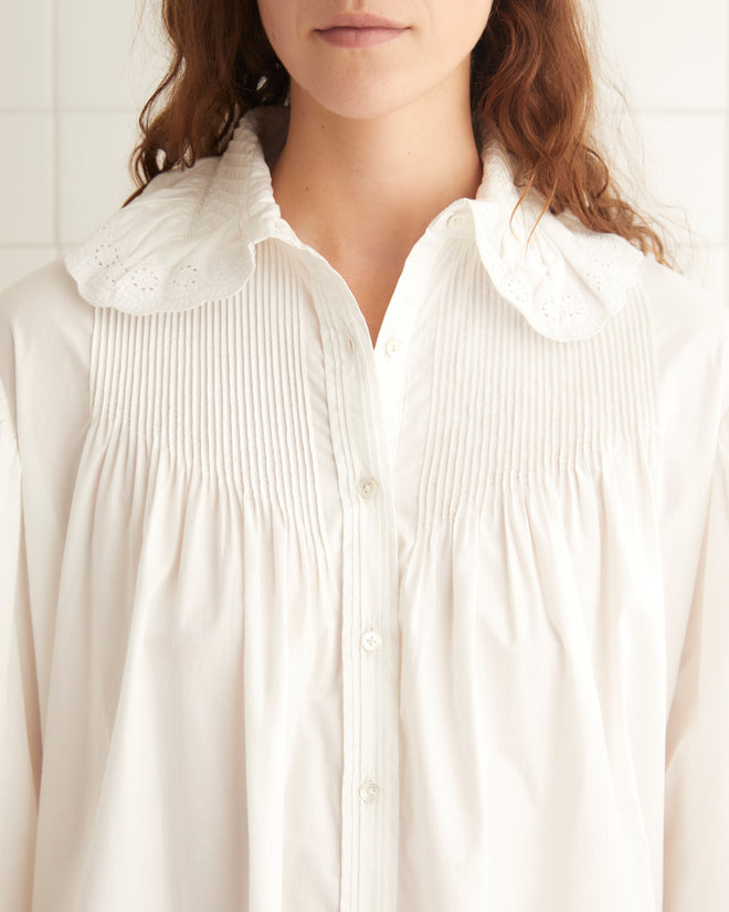 A person stands in a white tiled room wearing the Petaluma Dress in white, paired with black shoes.