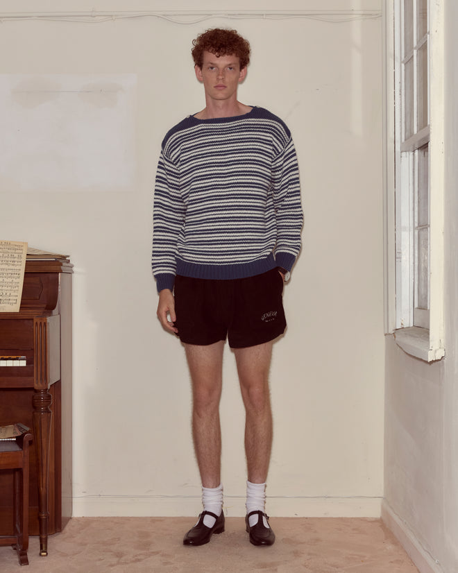 A young man in a blue and white Peter Stripe Sweater, black shorts, white socks, and black shoes stands indoors by a piano.