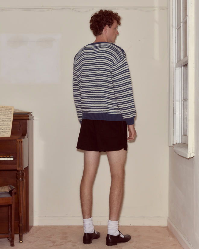 Wearing the Peter Stripe Sweater in blue and white, paired with black shorts, socks, and dress shoes, a person stands by a window facing a blank wall.