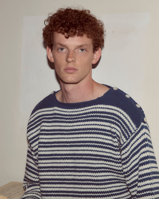 A young man with curly hair wears the Peter Stripe Sweater, a blue and white striped lambswool cashmere piece, looking directly at the camera.