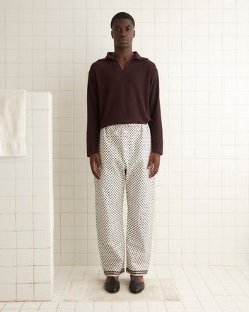 A man stands in a tiled room wearing a maroon long-sleeve shirt and the Petit Motifs Pajama Pant, which features a vintage-inspired black dot pattern. The 100% cotton pants, designed with a men's pajama silhouette in mind, are complemented by black shoes. A white towel hangs on a divider to his left.