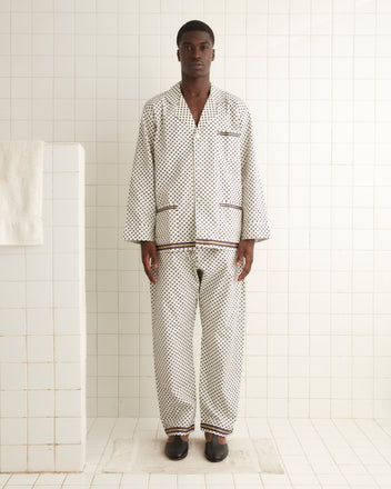 A person stands in a bathroom, wearing the Petit Motifs Pajama Shirt and black shoes, against a white tiled wall and floor. The men's pajama silhouette, complete with 100% cotton fabric, evokes a cozy yet timeless feel.