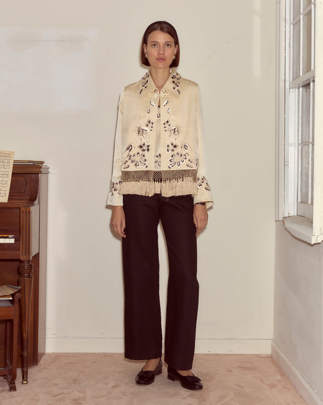 A woman stands indoors by a piano and window, wearing the Phantasm Fringe Jacket in ivory silk with Victorian embroidery, paired with black pants.