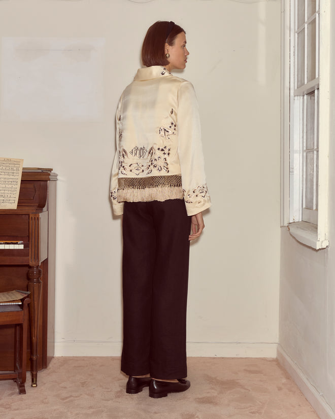 A woman stands indoors, facing away, wearing the ivory silk Phantasm Fringe Jacket with Victorian embroidery and black pants beside a piano and window.
