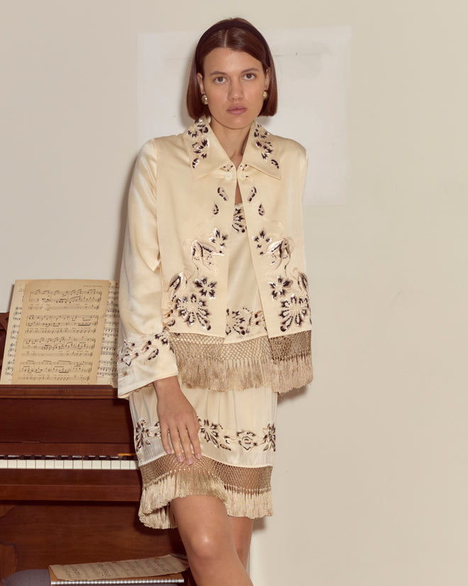 Woman in a cream embroidered outfit and the Phantasm Fringe Jacket stands by a piano with sheet music, looking at the camera.