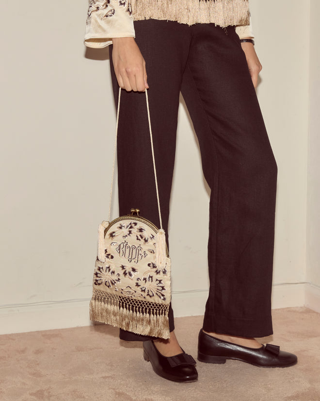 A person in black pants holds the Phantasm Fringe Bag—an ivory silk satin accessory with Victorian embroidery and fringe—while wearing black loafers.